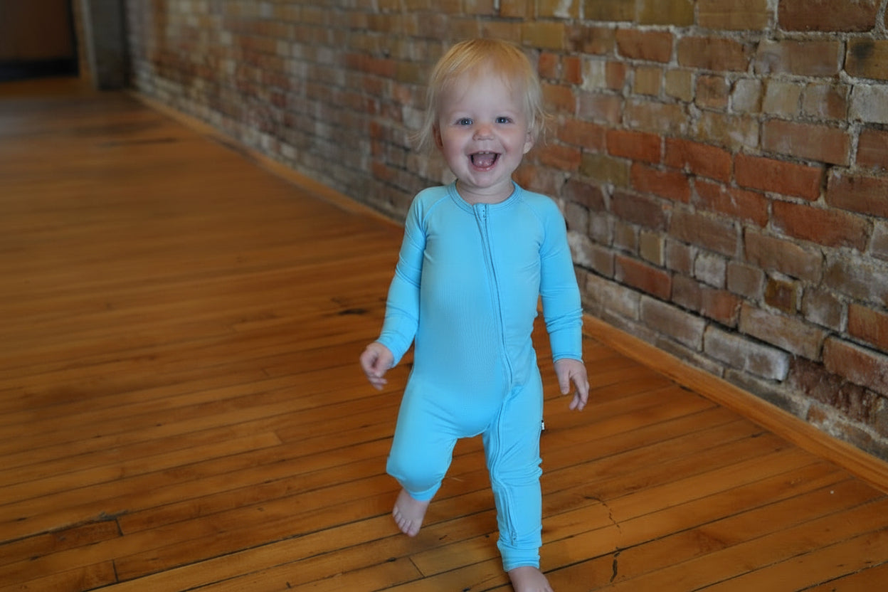 Child in a blue onesie standing on a wooden floor with a brick wall background